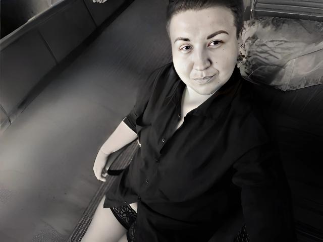 RachelExcite with light skin and dark hair, wearing a black shirt and black lace panties, sitting on a bed, smiling at the camera, showing a subtle pout.