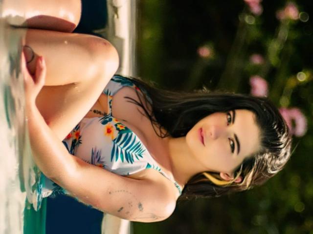 JeneKurz with wet black hair, wearing a floral bikini, sitting with knees up, showing small tits, and a tattoo on her arm, looking at the camera.