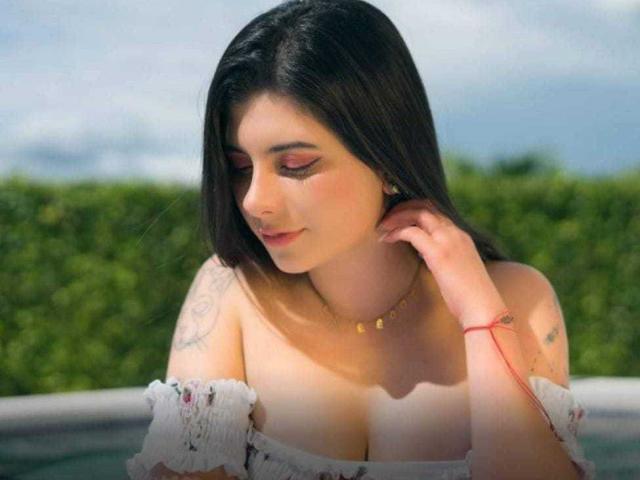 JeneKurz with dark hair, off-shoulder white top, showing big boobs, pink eyelids, gold necklace, tattoo on right shoulder, red bracelet, hand near neck, outdoor light.
