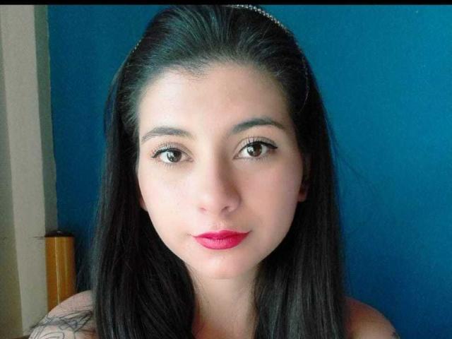 JeneKurz has long black hair, brown eyes, and red lipstick. She's wearing a silver headband, looking straight at the camera with a slight smile. Boobs are not visible.