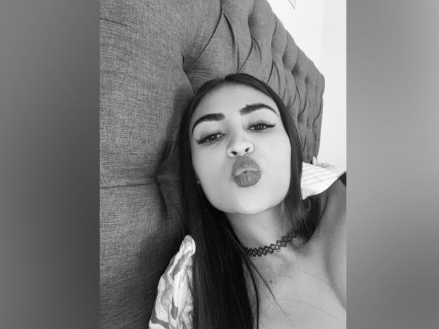 MiilaWx with pouty lips, thick eyebrows, long dark hair, choker, and gray upholstered headrest, looking up with a slightly open mouth. Boobs partially visible. Black-and-white photo.