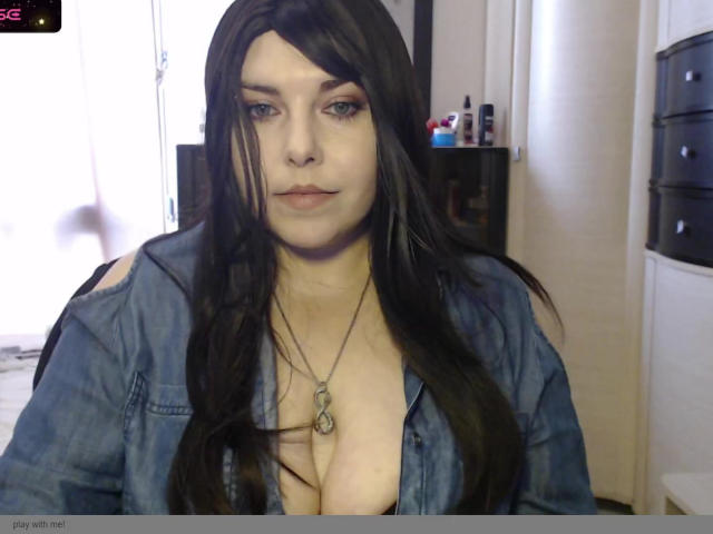 WhitteLotus with long black hair, blue eyes, and a low-cut denim shirt, showing her tits, wearing a necklace, sitting in a room, looking at the camera.