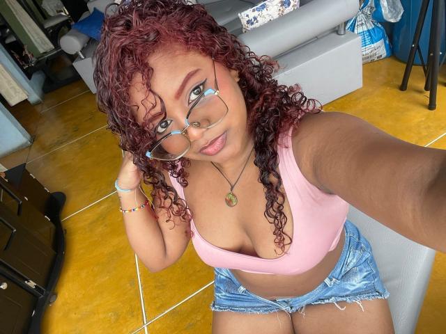 Ivonnerice with curly red hair, glasses, pink top, showing big tits, denim shorts, yellow floor, holding glasses, necklace, and bracelet, smiling at camera, sitting on chair.