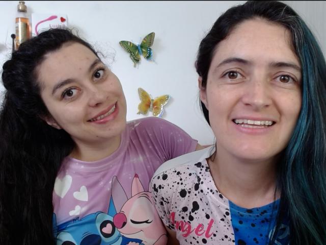 LunaVenus with dark curly hair in a pink heart shirt and LunaVenus with dark hair and blue-green highlights in a pink and black splatter shirt, smiling. Butterflies on wall.