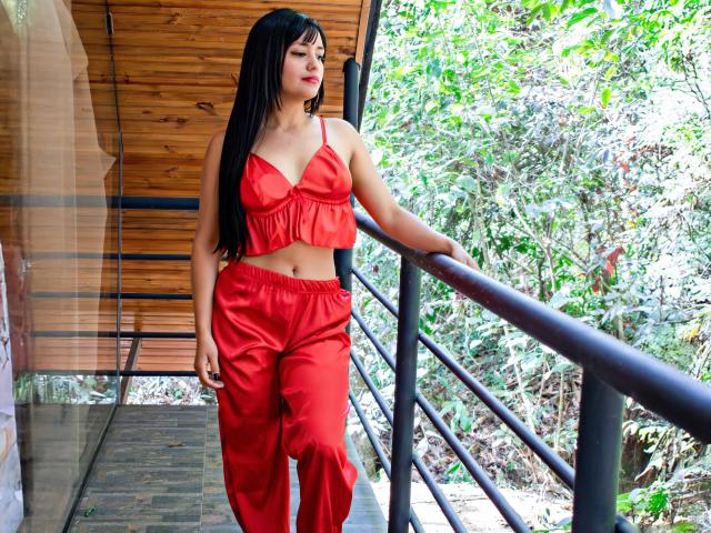 SilvannaReyes with long black hair, red crop top, and red pants, standing on a wooden balcony, leaning on a black railing, looking to the side with a slight smile.