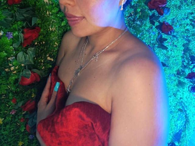 ArielleWilde with bare shoulders, red velvet fabric covering her tits, blue nail polish, silver necklace, and rose earrings, smiling slightly, green and red floral background.