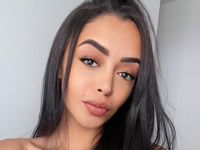 VicBrazilian has tan skin, thick black eyebrows, long straight black hair, brown eyes, and full pink lips. Her face is slightly tilted with a subtle, confident smile.
