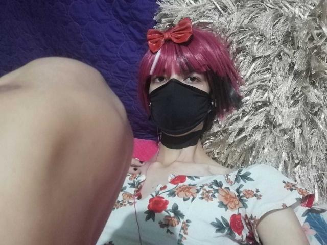 Tulipanti mit rosa und schwarzem Haarschopf, rotem Schleifchen, schwarzer Maske, Blumenkleid und freiem Bein, trägt ein rotes Schleifchen im Haar. Sie schaut nach oben mit einem niedlichen Ausdruck..