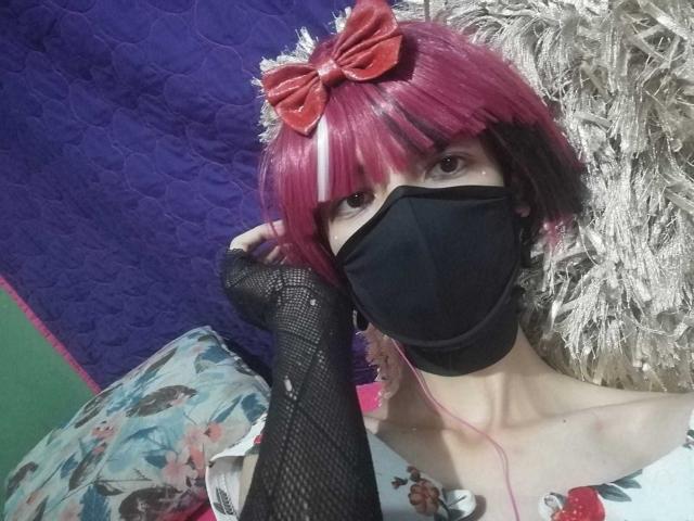 Eine Tulpenfigur mit rotem und schwarzem Hut, rotem Schleifchen, schwarzer Maske, Fischernetz-Arm, rosa Kopfhörern, die auf einem flauschigen Teppich liegt, trägt ein Blumenkleid, der Hintergrund ist grün und lila. Brüste sind teilweise sichtbar..