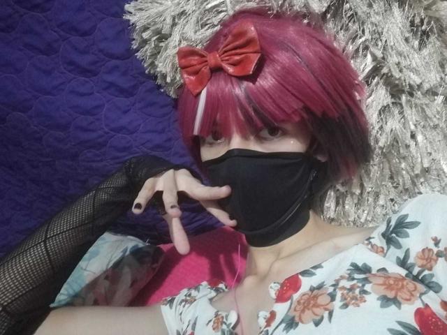 Tulipanti con flequillo rosa, lazo rojo, máscara negra, guantes de malla en el brazo, vestido floral, mostrando el dedo a la boca, uñas negras, piel pálida, acostada sobre un almohadón gris suave, colcha morada..