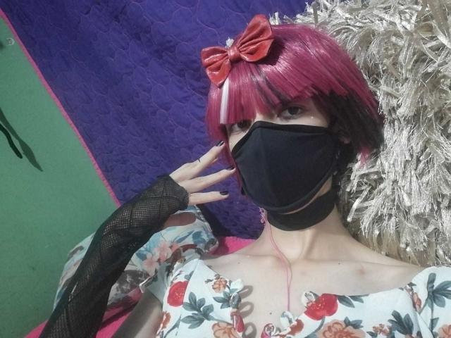 Tulipanti com um corte de cabelo rosa, um laço vermelho, uma máscara preta, um vestido floral, luvas pretas sem dedos, segurando o rosto. Fones de ouvido rosa. Pele clara, feminina. Uma parede branca e irregular ao fundo..