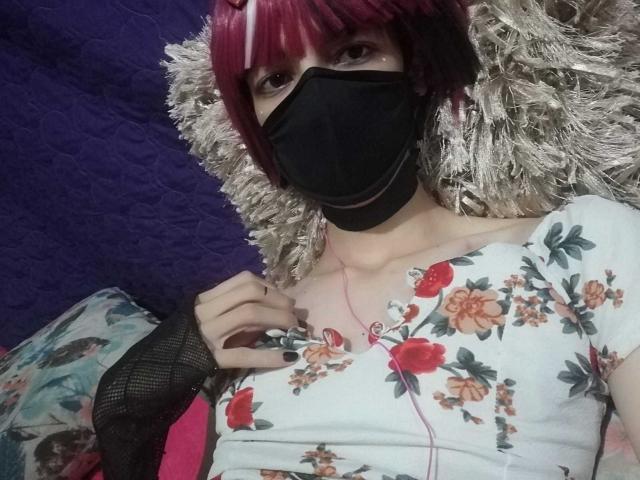 Tulipanti with red bob, black mask, floral top, and fingerless glove, lying on a fluffy pillow, pink earbuds visible, holding shirt, purple background, slightly visible ass.