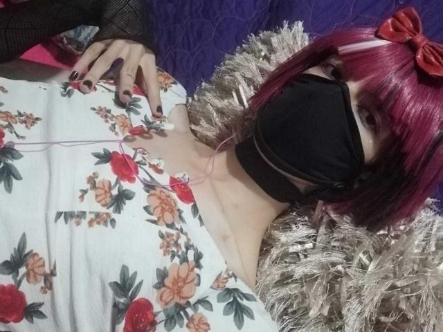 Eine Frau namens Tulipanti mit rot-pinken Haaren, einer schwarzen Maske, einem weißen Oberteil mit Blumenmuster, einer schwarzen Halskette, einem Netzarmstulpen und liegt auf einem beigen Fell. Ihre Hand liegt auf ihrer Brust, im Hintergrund ist Lila zu sehen, und sie trägt Kopfhörer. Ihre Brüste sind sichtbar..