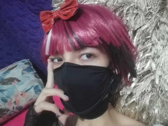 Eine Tulpe mit rosa und schwarzen Haaren, einem roten Schleifchen, einer schwarzen Maske, einem Finger, der die Maske berührt, die in die Kamera schaut und einen schwarzen Handschuh ohne Finger trägt. Im Hintergrund ist eine silbrig strukturierte Wand zu sehen..