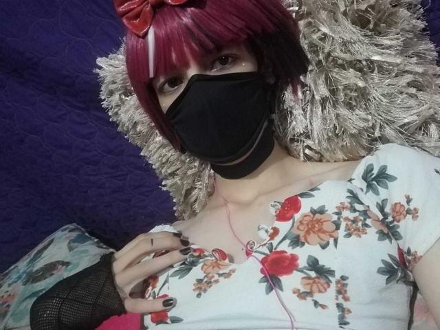 Tulipanti with red bob, black mask, floral top, and black finger mesh, lying on silver tinsel, looking at camera. Boobs visible, pink earbuds, black nail polish, tattoo on finger.
