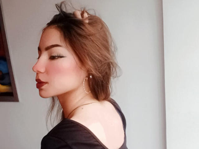 AshleyMiler with brown hair, hoop earrings, and black top, side profile, holding hair, eyeliner, pink blush, and red lipstick, showing a tan line on her back.
