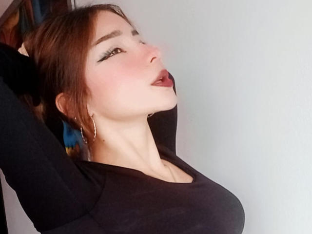 AshleyMiler has her arms raised, showing off her tits in a black top, with her mouth slightly open and tongue out, wearing hoop earrings, and looking up.