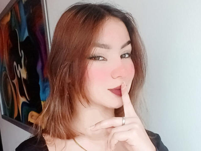 AshleyMiler with pink blush, brown eyes, and dark lipstick, touching her finger to her mouth, showing her ring. She has shoulder-length brown hair and a black top.