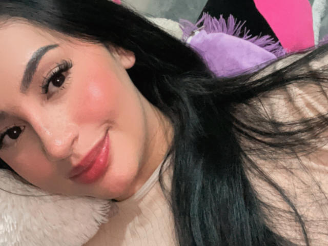 MartinaMendezz with long black hair, red lipstick, and dark eyebrows, smiling at the camera. Her bare shoulders are visible. She has a cute, sexy look.