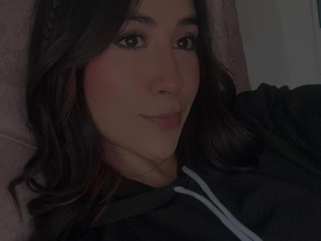 AnyaJay with dark brown wavy hair, subtle pink blush, and black hoodie, smiling slightly with soft pink lips. Dark eyes, light skin, and relaxed pose.