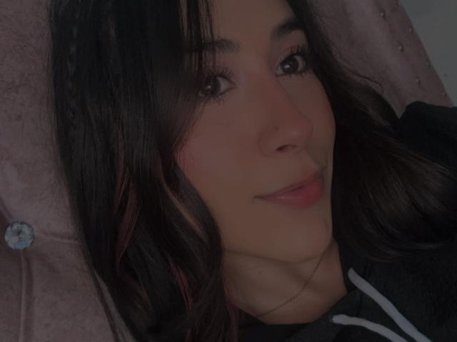 AnyaJay with dark wavy hair, brown eyes, and a slight smile, lying down with a pink tufted pillow behind her head, wearing a black hoodie with white stripes.
