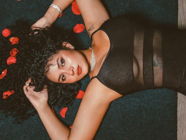 CanelaLebrad with curly black hair, wearing a black sheer dress, lying on dark green carpet, rose petals around her head, hands above, looking at camera, red lipstick, necklace.
