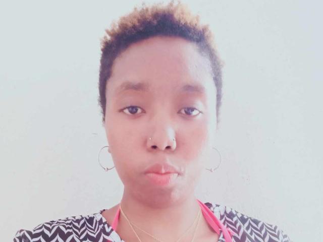 Jobba has short curly brown hair, a nose piercing, hoop earrings, and is pouting her lips while wearing a black and white zigzag patterned top with pink underneath.