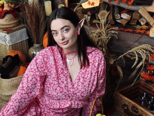 MilanaPrice with elf ears, pink floral dress, pink earrings, necklace, and cute smile, showing her tits in a cozy, autumnal setting with baskets, pumpkins, and fall decor.