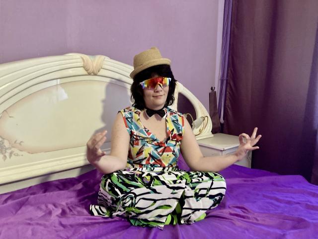 EverlyRays is sitting on a bed with purple sheets, wearing a colorful patterned dress, zebra pants, reflective glasses, a straw hat, and a black choker, meditating.