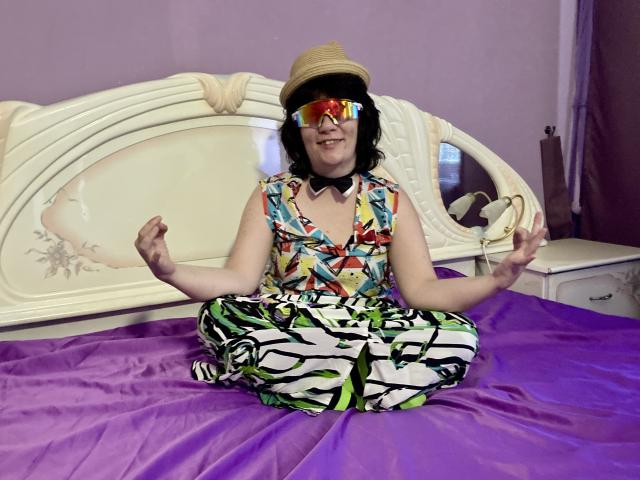 EverlyRays, wearing a straw hat, colorful sleeveless top, black bowtie, and reflective goggles, sits cross-legged on a purple bed, hands in a meditation pose, smiling.