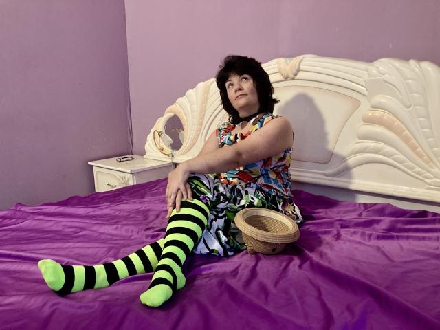 EverlyRays with black hair, colorful dress, and neon green and black striped socks, sits on purple bed, hand on wicker basket, looking up, in pink-walled bedroom.