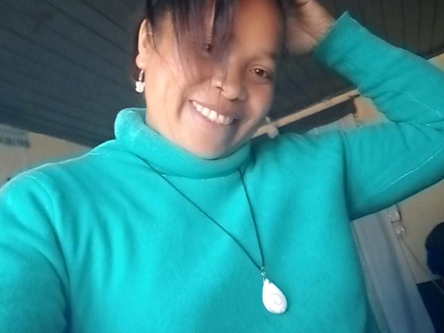 Rahel69 with short black hair, smiling, wearing a turquoise turtleneck, necklace with white pendant, and earrings, hand in hair, showing small nose ring and white teeth.