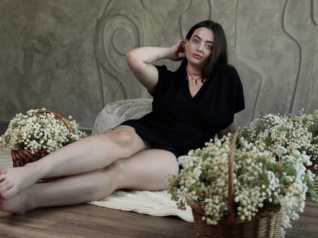 MilanaPrice with pale skin, dark hair, black dress, legs spread, sitting on rug, surrounded by baskets of white baby's breath, hand on head, pink necklace, subtle tattoo.