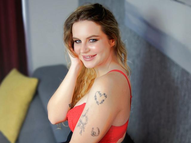 ZoeyFancy with wavy blonde hair, red lace bra, smiling at camera, hand in hair, tattoos on right arm, blue eyes, cute, soft light, indoors, sexy.