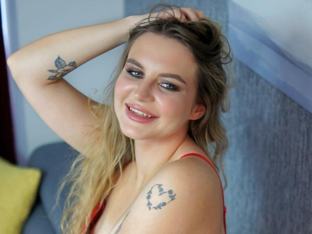 ZoeyFancy mit blonden Haaren, blauen Augen und einem roten Tanktop, lächelt in die Kamera, hat eine Hand im Haar, Tattoos am Arm und an der Schulter, ist süß, attraktiv, hat kleine Brüste, rosafarbene Lippen, lässig..