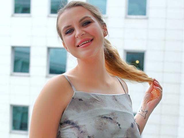 ZoeyFancy has light skin, blue eyes, and a smile showing teeth. She's wearing a gray tie-dye top, holding her blonde ponytail, with a tattoo on her right wrist.