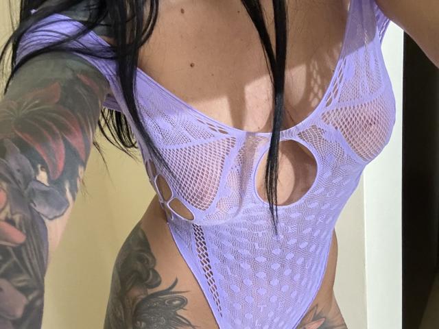 AndraD wearing a purple mesh bodysuit with cutouts, showing her tits and tattoos on her arms and thighs. Her black hair is down, partially covering her face.