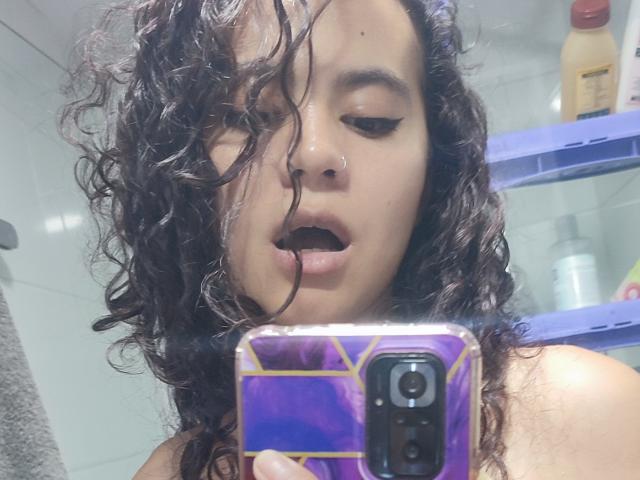 SuzzyV with curly black hair, wet face, nose ring, and open mouth, holding a purple phone with gold lines, in a bathroom, looking at the camera.