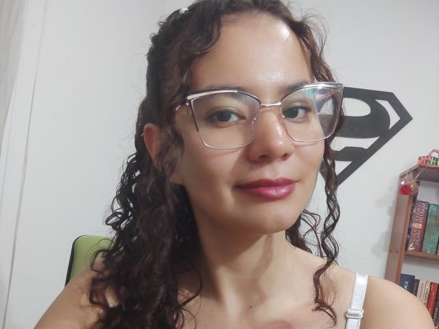 SuzzieV has curly brown hair, glasses, and pink lipstick. She's smiling with shiny skin, wearing a white strap top. Her tits are small, and she's cute.