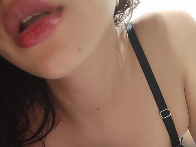 SuzzieV's face, showing red lipstick, slightly open mouth, black strap, bare collarbone with a small mole, and curly dark hair, focusing on her upper chest and lips.