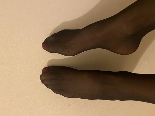 Dionisia's feet in sheer black pantyhose with red-painted toenails, toes pointing slightly upward, legs positioned vertically against a plain wall.