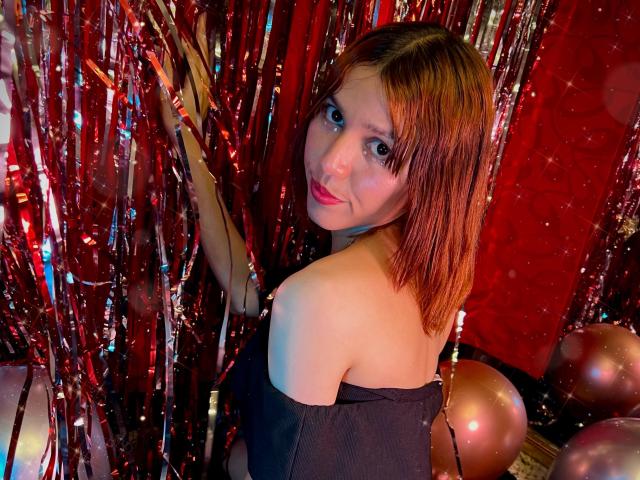 RachelJonier with red-brown hair, looking over her shoulder, wearing an off-shoulder black top, standing in front of shiny red and silver metallic strips, with pink balloons.