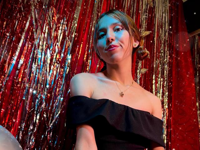 RachelJonier with red and gold metallic curtain, wearing black off-shoulder dress, small tits, brown hair in bun, smiling, gold necklace, red light, soft blue light on face.