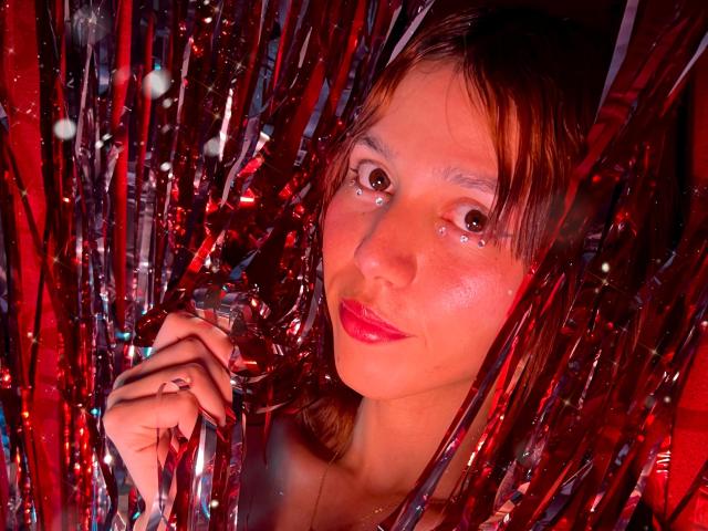 RachelJonier with shiny red lips, brown eyes, and wet-looking skin, holding metallic red and silver strips, giving a sultry, serious look. She has a small nose and brown hair.