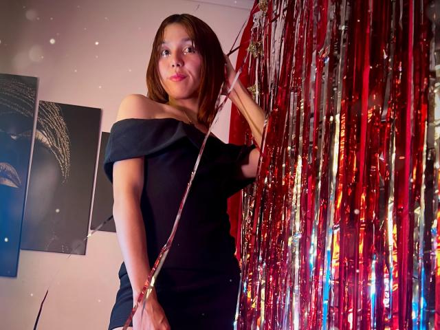 RachelJonier with medium tits, brown hair, black off-shoulder dress, holding red metallic streamers, standing slightly to the right, looking up with a subtle smile.