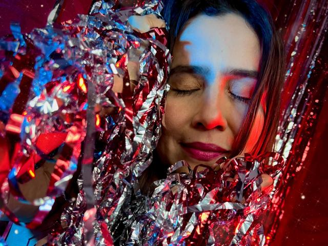 HeidyWells with shiny silver tinsel covering her face and arms, eyes closed, red and blue lights on her skin, wearing pink lipstick, looking relaxed and happy.