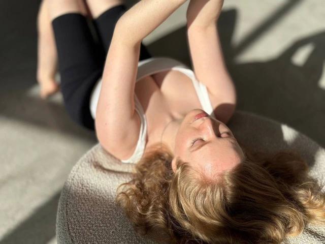 SunnyBunny-hot with curly blonde hair, lying on a textured gray surface, wearing a white tank top and black pants, sunlit, eyes closed, arms raised.