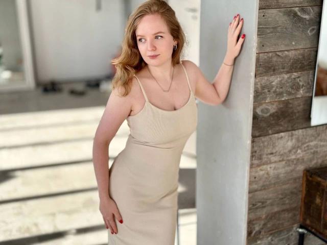 SunnyBunny-hot with pale skin, blonde hair, and red nails in a beige dress, standing against a wooden wall, hand on surface, looking at camera, small tits, cute smile.