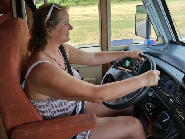 Jutta1-hot with curly brown hair, white headband, tank top, and checkered dress, driving, holding steering wheel, smiling, tan car seat, visible green car interior, daytime.