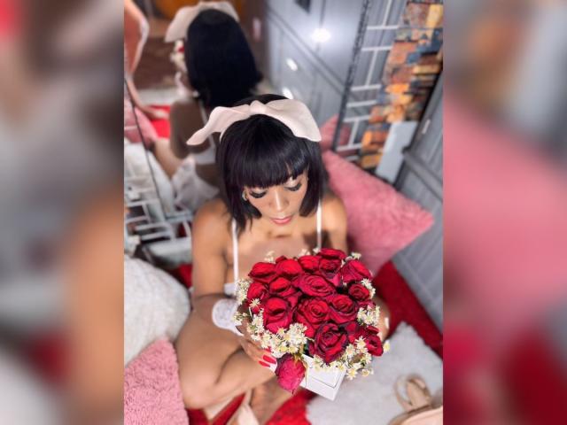 GinaChaudeX with short black hair, white bunny ears, and red lipstick, holding a bouquet of red roses and white flowers, sitting on a pink and red carpet.